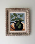 A framed still life painting featuring a black pitcher with green leaves on a light blue and brown background, housed in a distressed, ornate textured frame. Available at Hue.