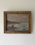 A framed vintage coastal landscape oil paintingl. The artwork depicts rocky shoreline, ocean waves, distant hills, and a cloudy sky, enclosed in an ornate textured gold-toned frame. Available at Hue.