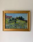 A framed vintage landscape oil painting featuring green fields, trees, and a red-roofed building under a blue sky, housed in a distressed gold frame. Available at Hue.