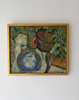 A framed vintage still life oil painting depicting a blue patterned plate, a basket of fruit, and bottles on a draped tablecloth. Available at Hue.