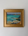 A framed vintage coastal landscape painting with turquoise water, rocky shoreline, and distant hills, enclosed in a gold frame. Available at hue.