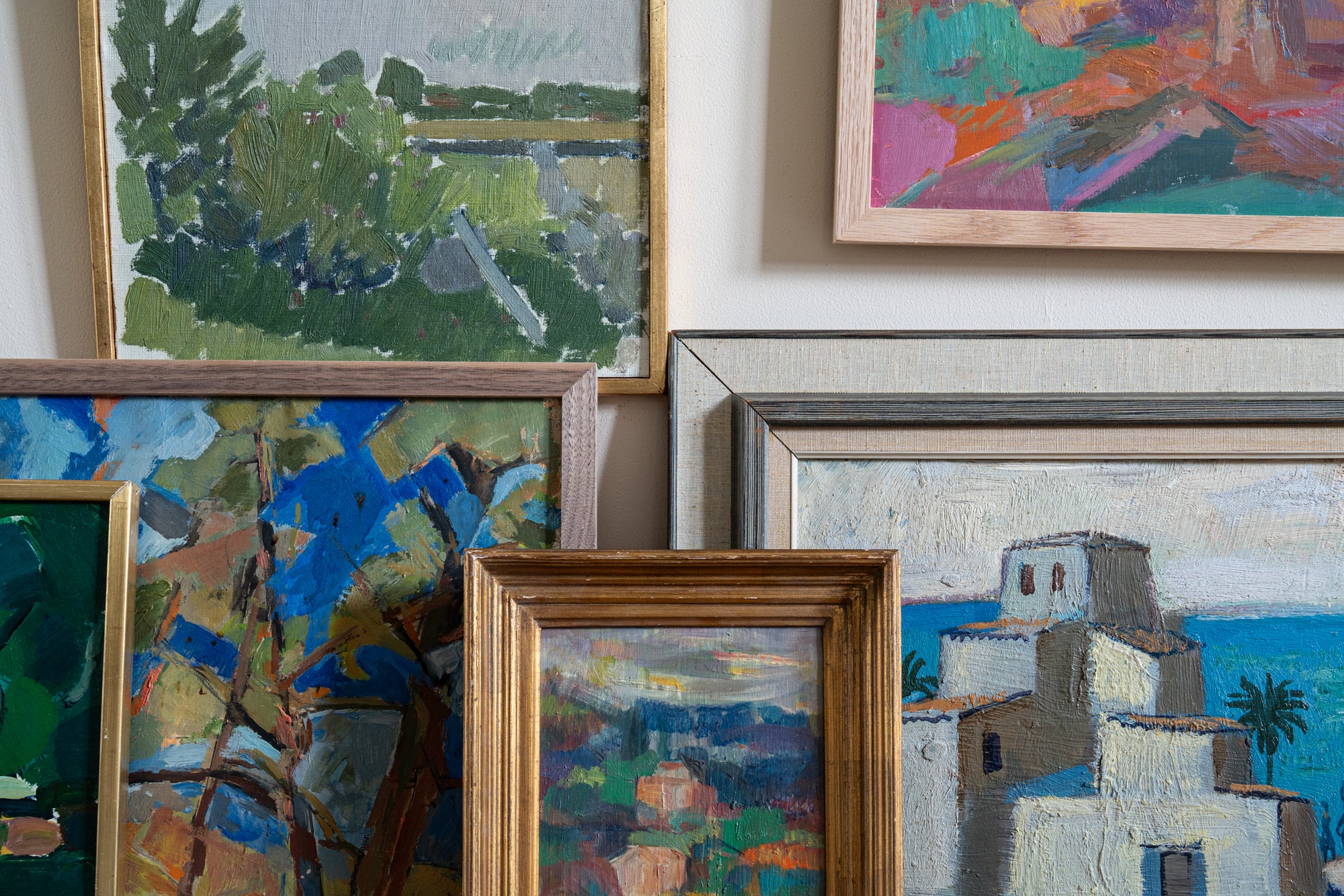 Collection of framed paintings on a wall with some propped up, colourful mix with blues, greens and pinks