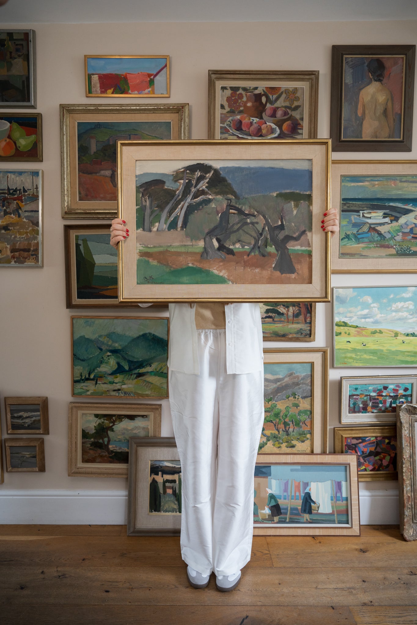 Person holding a framed painting in front of a wall of other framed artworks.