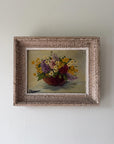 A vintage framed still life painting of a colorful flower bouquet in a dark red bowl, set against a neutral beige background. Available at Hue.