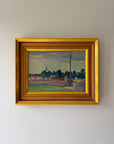 A framed vintage landscape oil painting. The painting depicts a rural scene with green fields, dark trees, and a cloudy sky, enclosed in a wide gold frame.