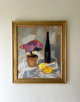 Framed vintage still life painting with a bottle, potted pink roses, and lemons on a table, available at Hue.