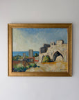 A framed vintage oil painting depicting a coastal landscape with white stone buildings, a blue sea, and a pale sky, housed in a gold-toned frame. Available at Hue.