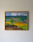 A framed vintage landscape oil painting. The painting depicts rolling green hills, yellow fields, a brown foreground, and a blue-gray cloudy sky. Available at Hue.