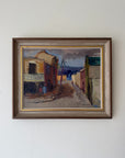 A framed vintage painting of a narrow street with rustic buildings and a palm tree.The artwork has earthy tones with blue, brown, beige, and muted red accents, enclosed in a dark brown wooden frame with a gold inner border. Available at Hue.