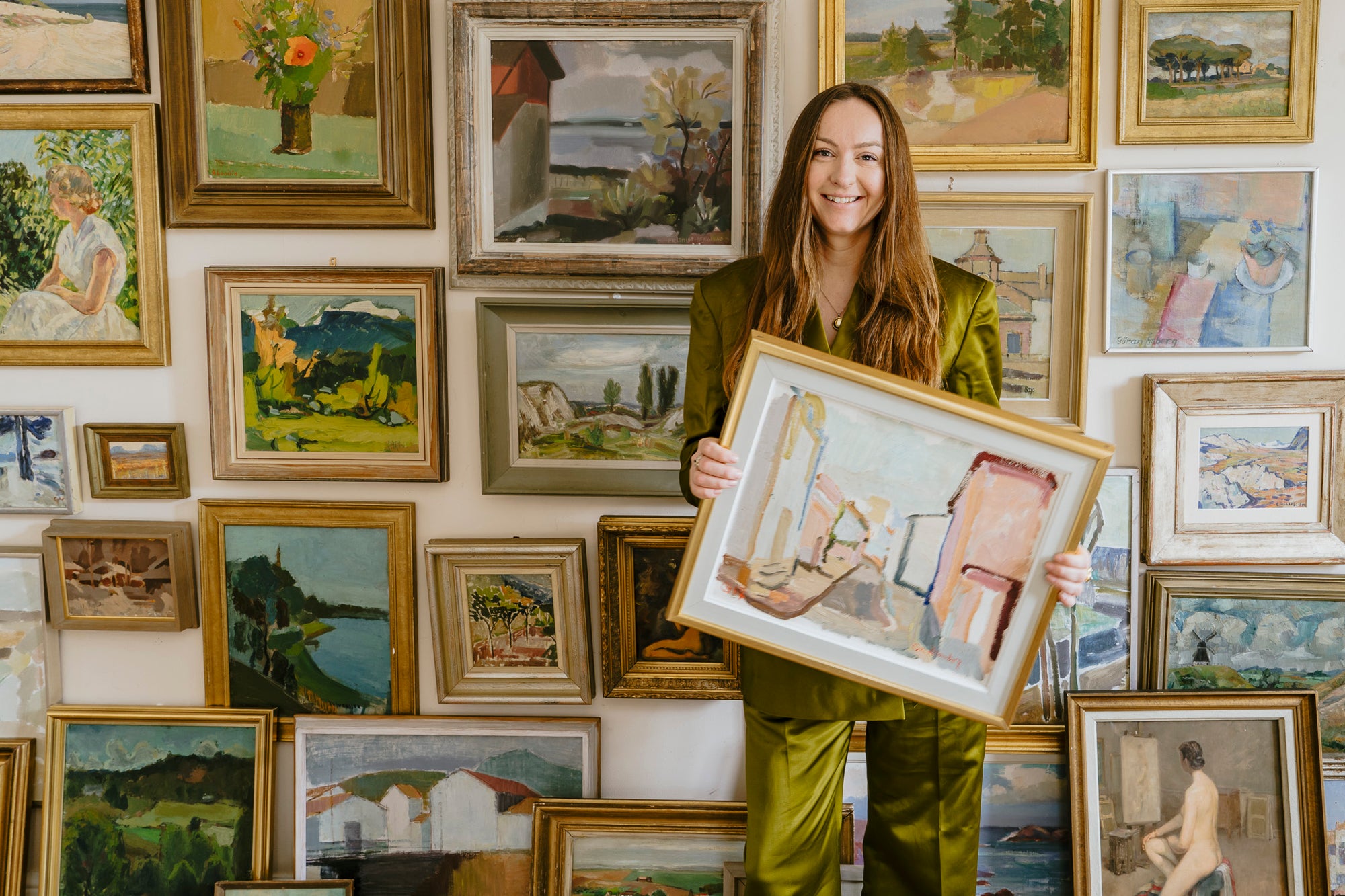 Woman holding a framed vintage painting in front of a wall of framed vintage artworks