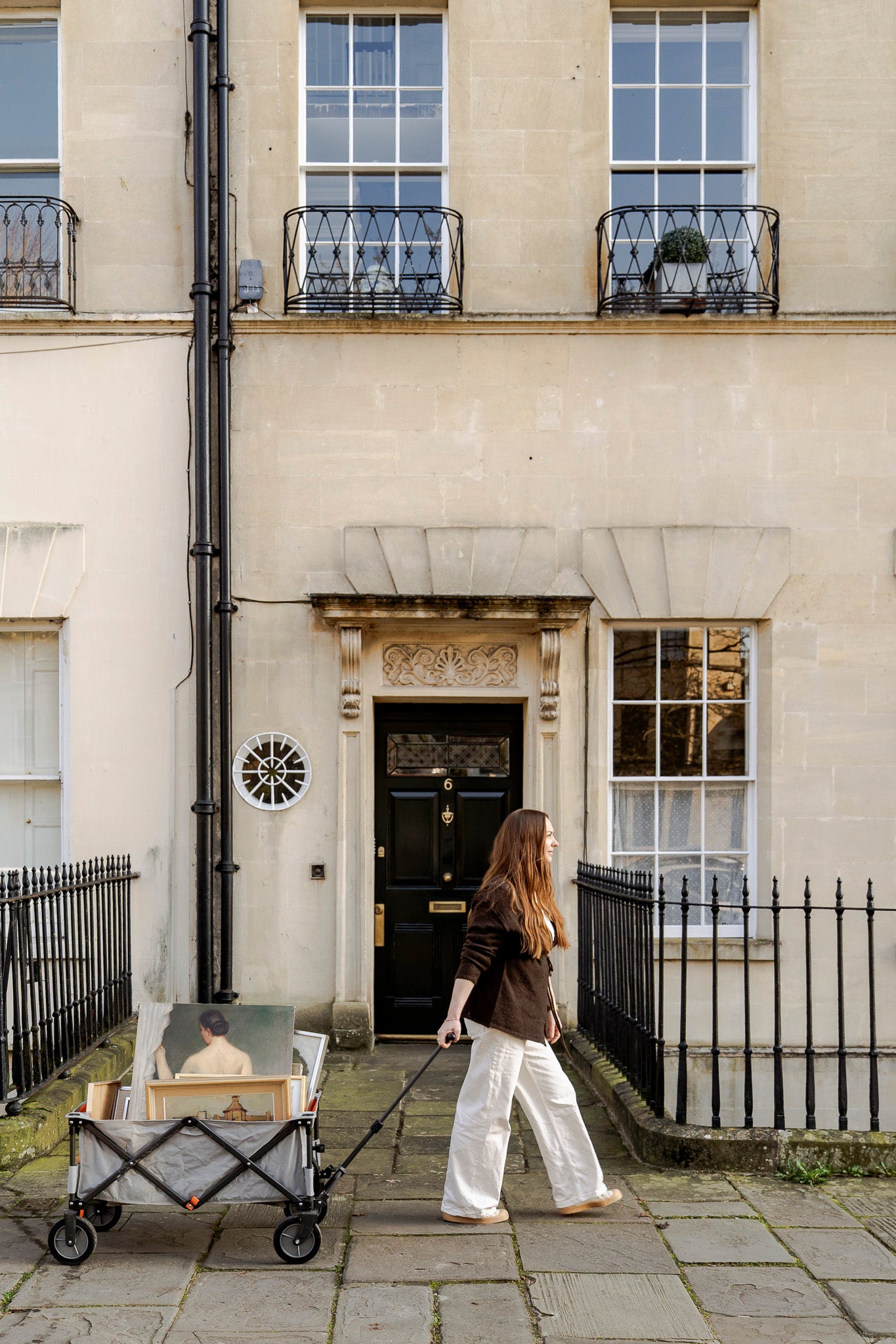 Woman walking past a large bath stone building with a black door and decorative elements pulling a trolley of vintage paintings