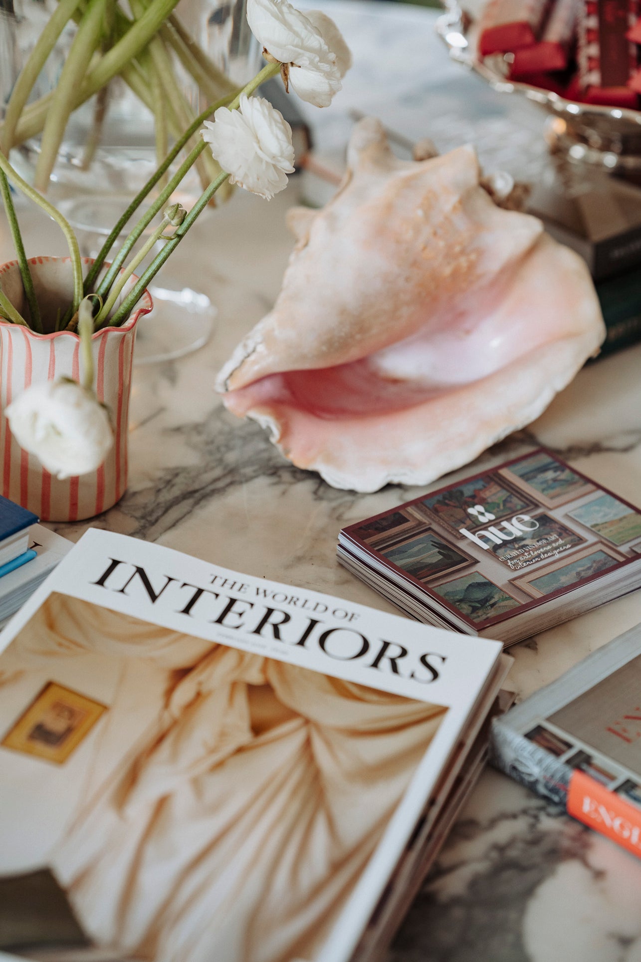 Book titled 'The World of Interiors' on a marble surface with decorative items including a shell and flowers and hue branded cards