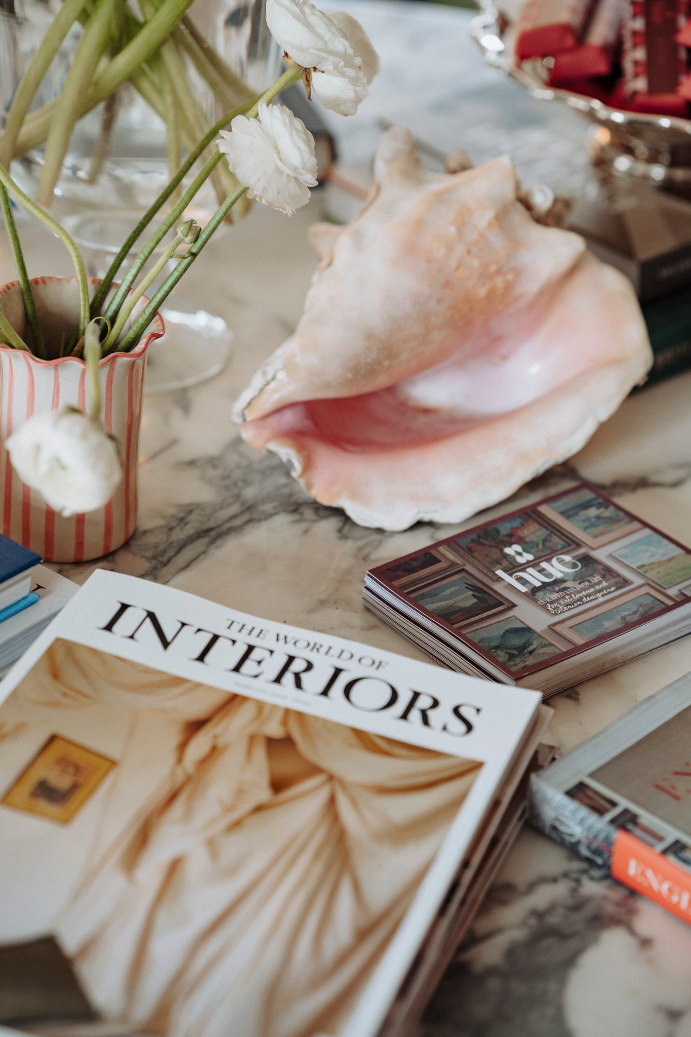 Book titled 'The World of Interiors' on a marble surface with decorative items including a shell and flowers and hue branded cards