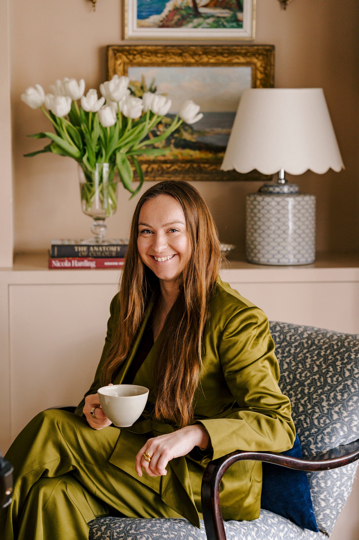 Woman in a green outfit holding a white mug in a cozy room with a lamp, paintings and flowers.