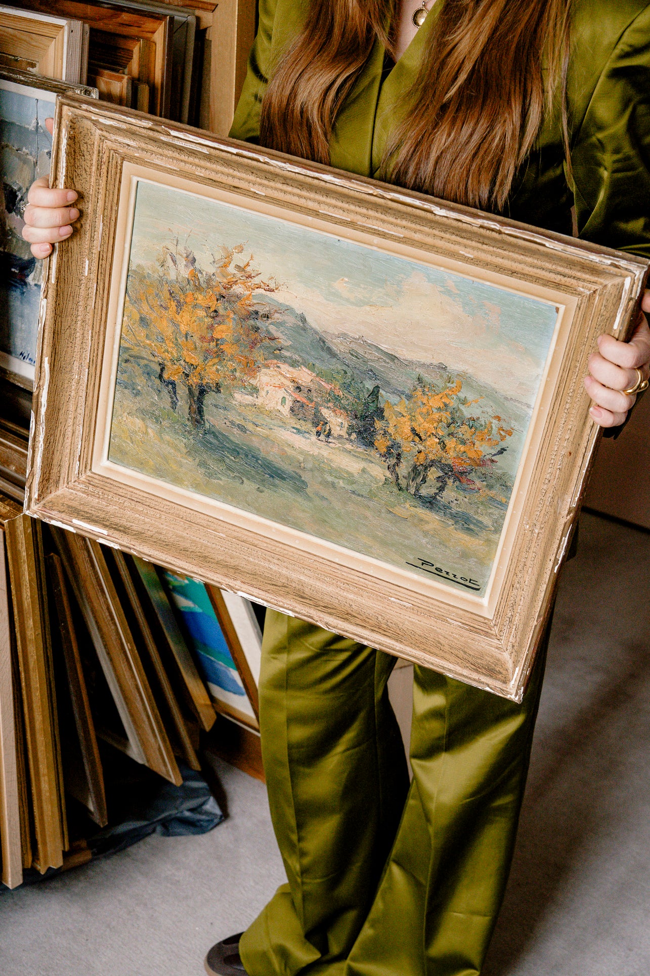 Person holding a framed painting of a landscape with trees and a house.
