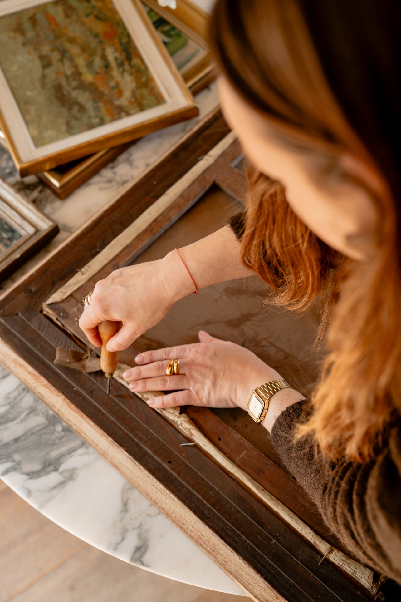 Person working on a wooden frame with a tool in a setting with framed artworks.