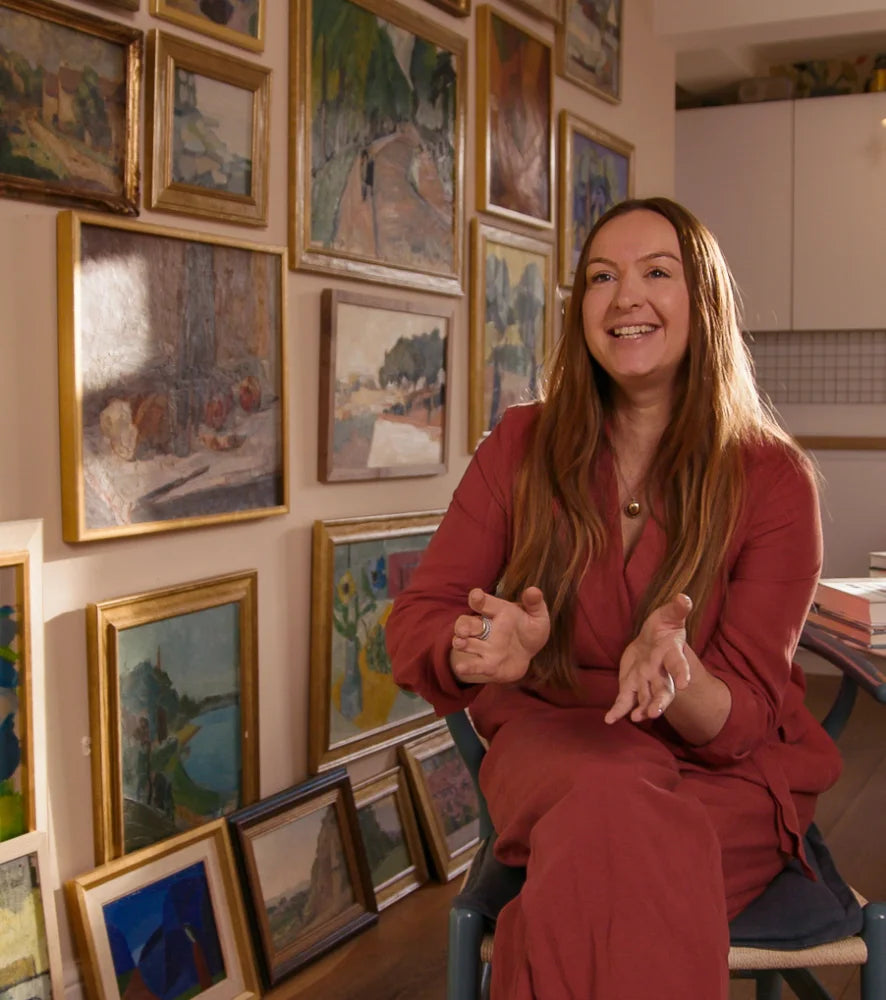 Hue + Holasz founder seated at home surrounded by a collection of framed vintage oil paintings.