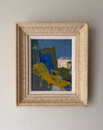A framed abstract painting featuring a bright yellow lounge chair against a blue and green background, housed in a textured cream-colored frame. Available at Hue. 