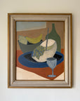 A framed still life painting. The artwork features a stylized arrangement with a blue plate, a white fruit, green grapes, a green bottle, and a blue glass, enclosed in a light wood frame. Available at Hue. 