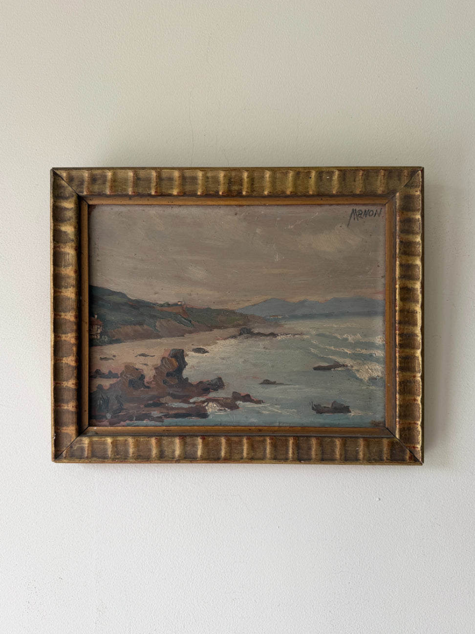 A framed vintage coastal landscape oil paintingl. The artwork depicts rocky shoreline, ocean waves, distant hills, and a cloudy sky, enclosed in an ornate textured gold-toned frame. Available at Hue. 