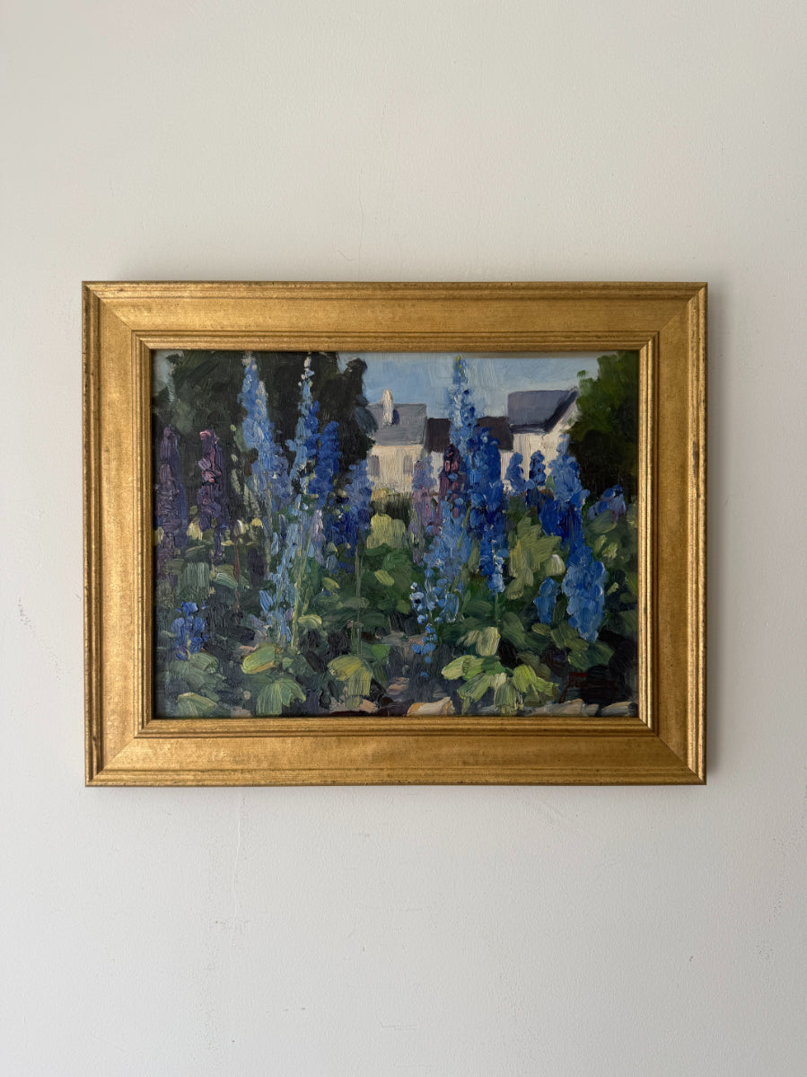 A framed vintage landscape oil painting featuring blue delphiniums and green foliage with a white house in the background, enclosed in a gold frame. Available at Hue. 