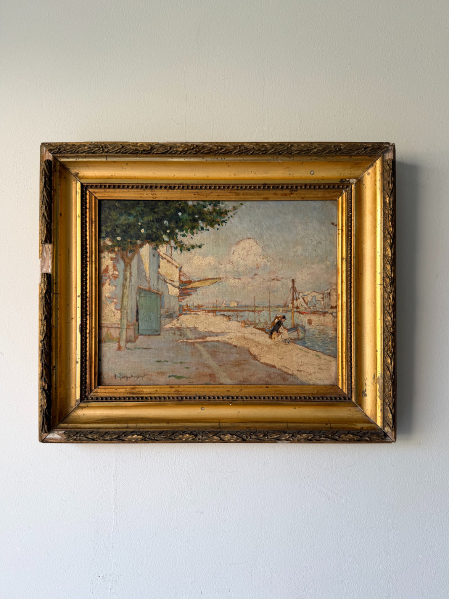 A framed vintage oil painting depicting a coastal street scene with a blue door, trees, and a harbour, housed in an ornate gold frame. Available at Hue.