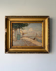 A framed vintage oil painting depicting a coastal street scene with a blue door, trees, and a harbour, housed in an ornate gold frame. Available at Hue.