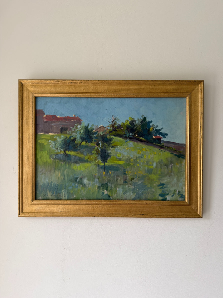 A framed vintage landscape oil painting featuring green fields, trees, and a red-roofed building under a blue sky, housed in a distressed gold frame. Available at Hue. 