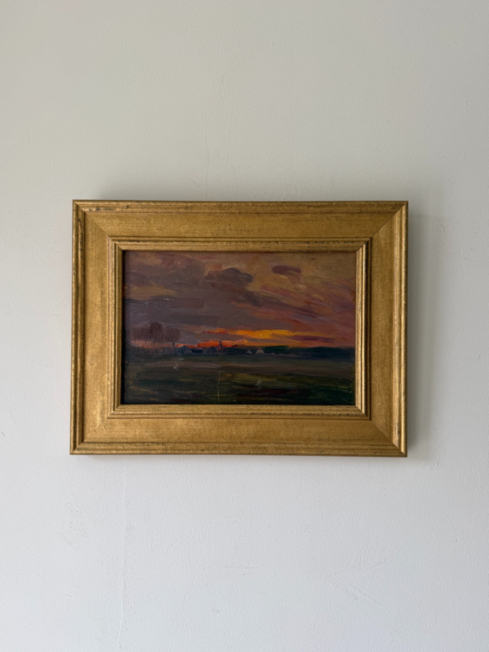 A framed vintage landscape oil painting. The painting depicts a sunset over a field with a dark, moody sky in shades of purple, orange, and brown, enclosed in a gold frame. Available at Hue.