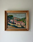A framed vintage oil painting of a Mediterranean village landscape, featuring a rustic stone building with red-tiled roofs, green hills, and a blue sky, housed in a distressed gold frame. Available at Hue. 