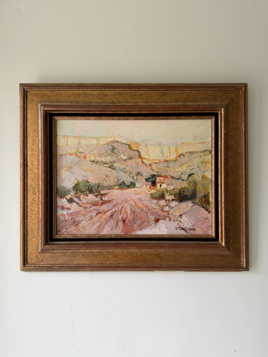 A framed vintage landscape oil painting The painting depicts a dusty, sunlit hillside village with mountains in the background, rendered in earthy pink, brown, and green tones. Available at Hue. 