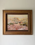 A framed vintage landscape oil painting The painting depicts a dusty, sunlit hillside village with mountains in the background, rendered in earthy pink, brown, and green tones. Available at Hue. 