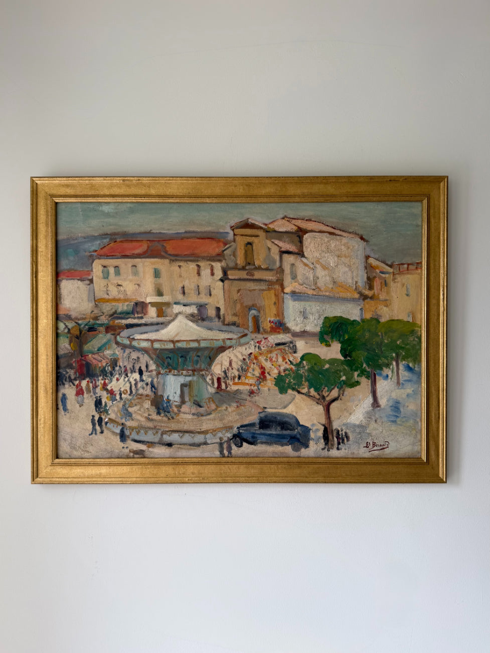 A framed vintage impressionist oil painting of a French town square, featuring buildings with terracotta roofs, market stalls, trees, and a dark car, enclosed in a gold frame. Available at Hue. 