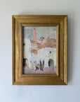 A framed vintage painting depicting a rustic village scene with white buildings, terracotta roofs, and small figures, housed in a wide gold frame. Available at Hue. 