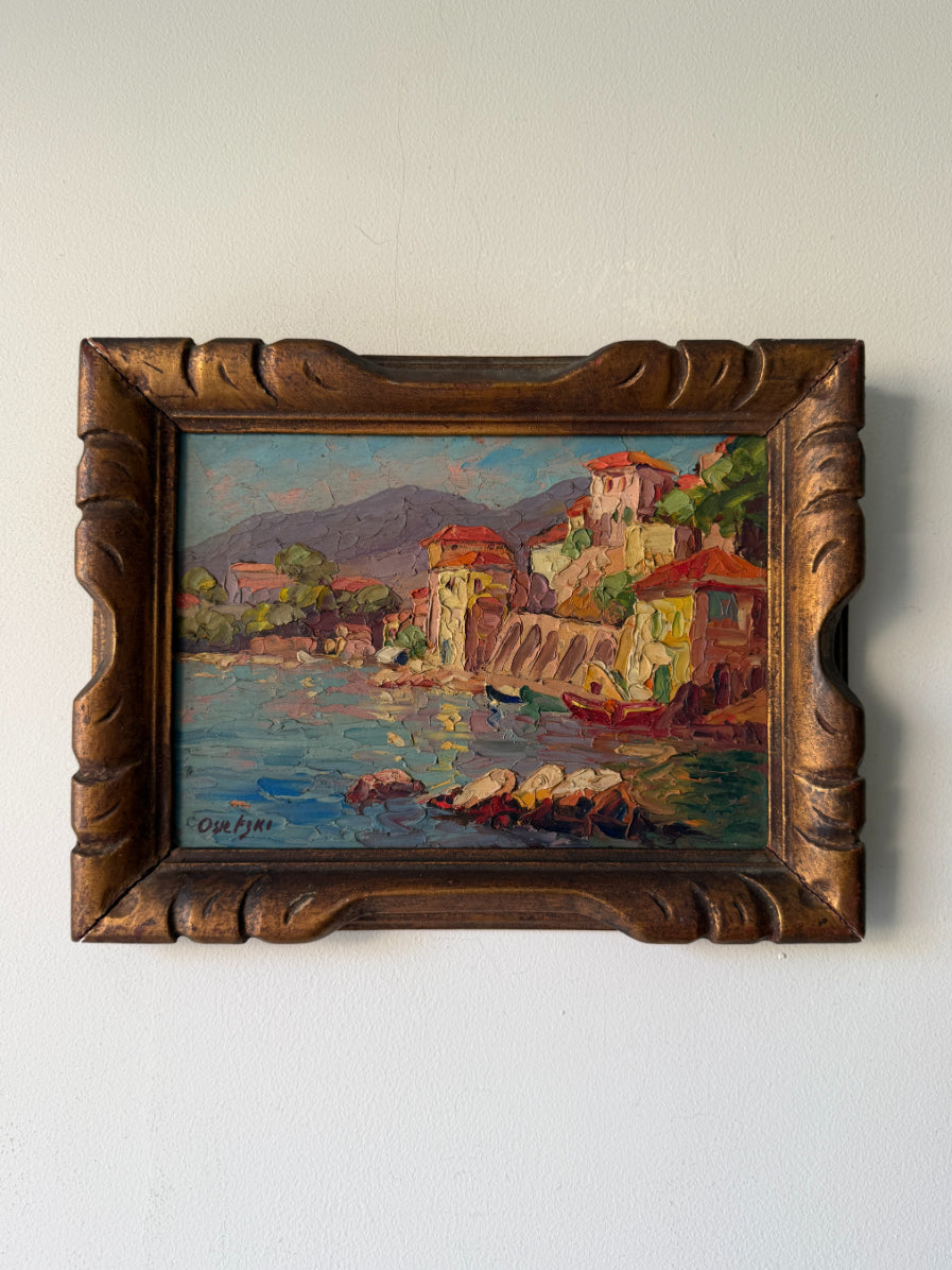 A framed vintage oil painting of a coastal village scene with colorful buildings, boats, and mountains, housed in an ornate carved wooden frame. Available at Hue.