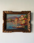 A framed vintage oil painting of a coastal village scene with colorful buildings, boats, and mountains, housed in an ornate carved wooden frame. Available at Hue.