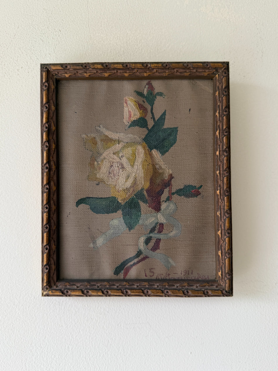 A framed vintage oil painting depicting a pale yellow rose with green leaves and a white ribbon, set in an ornate dark brown wooden frame. Available at Hue. 
