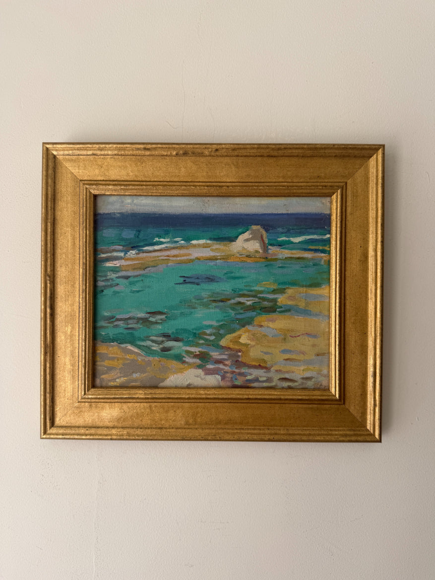 A framed vintage coastal landscape painting featuring turquoise water, rocky shoreline, and a dark blue horizon, housed in a wide ornate gold frame. Available at Hue. 