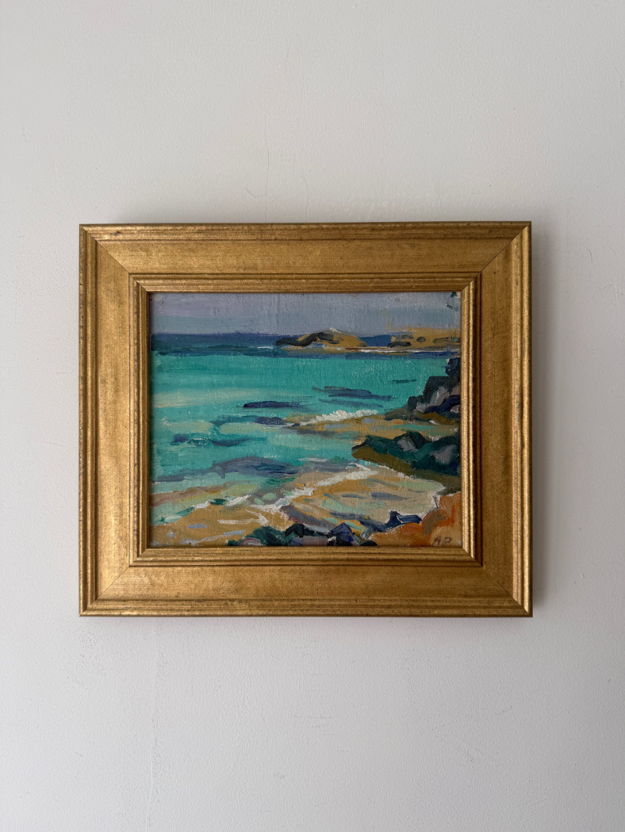 A framed vintage coastal landscape painting with turquoise water, rocky shoreline, and distant hills, enclosed in a gold frame. Available at hue. 