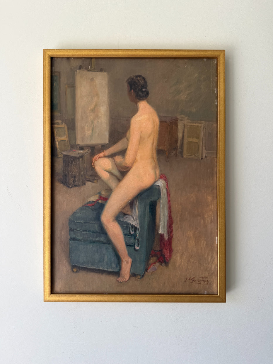 A framed vintage oil painting depicting a nude woman seated on a blue armchair in an artist studio, with a canvas and easel in the background. Available at Hue. 