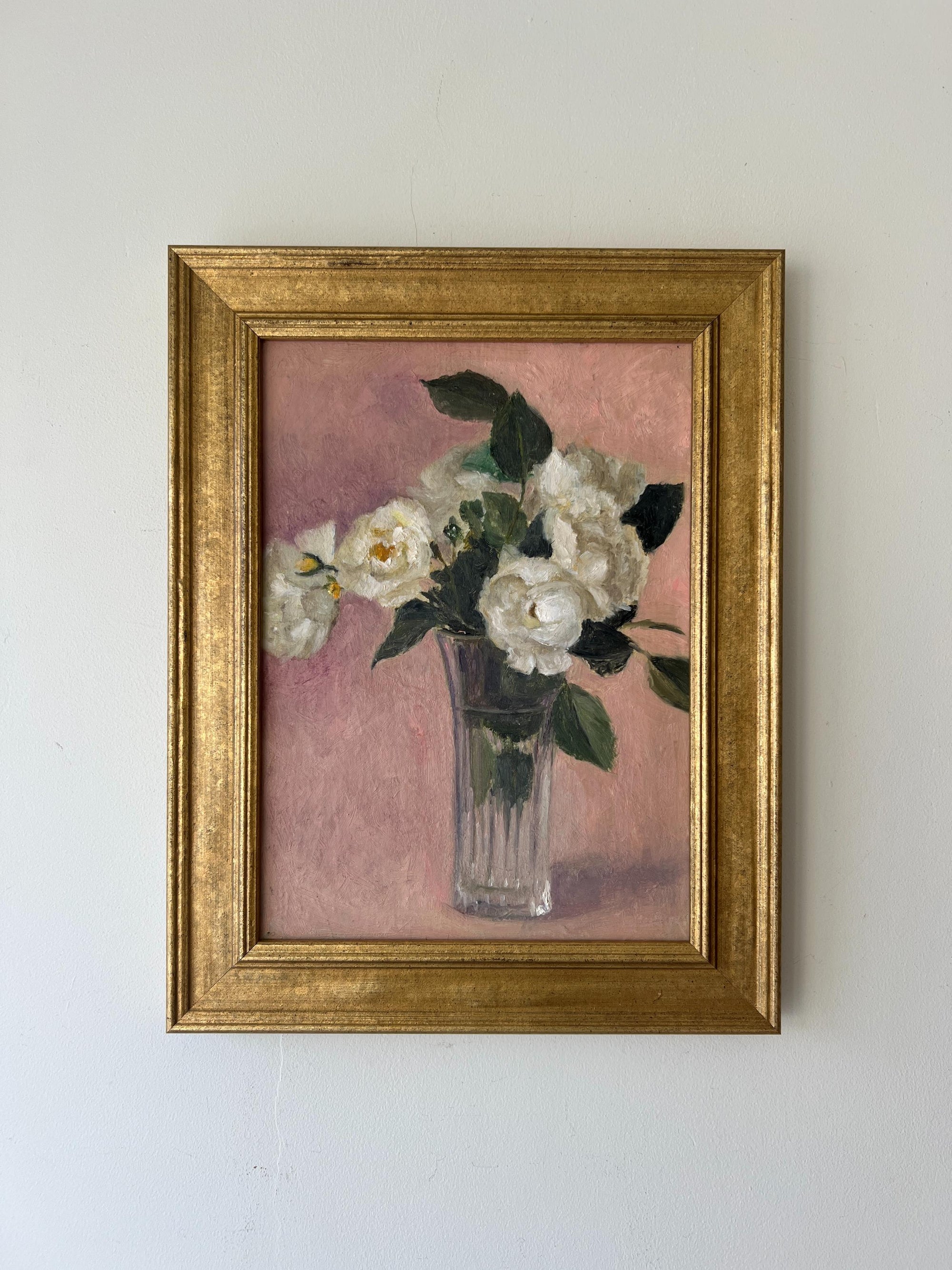 A framed vintage oil painting on canvas of a bouquet of white roses in a glass vase on a dusty pink background. Available at Hue.