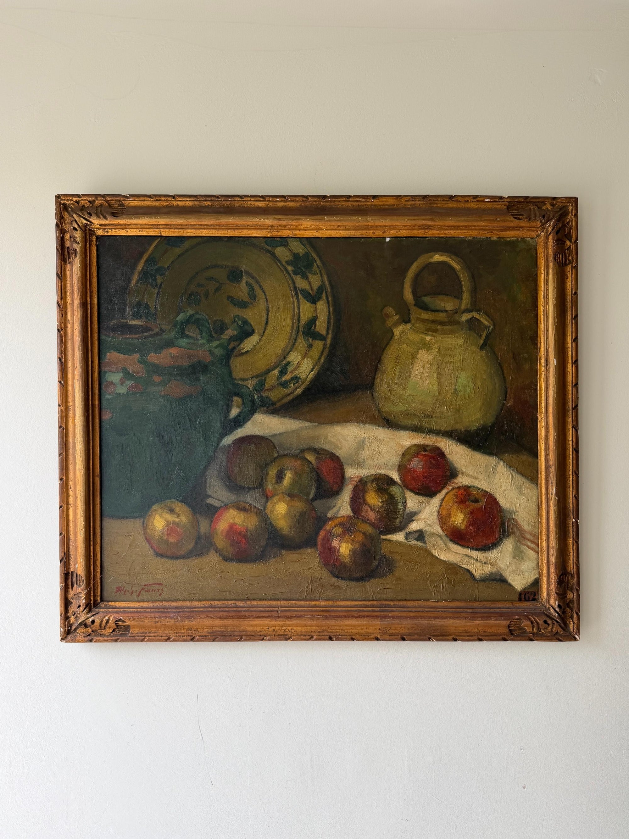 A framed vintage still life oil painting depicting apples, a green jug, a patterned plate, and a ceramic pot on a draped cloth. Available at Hue. 