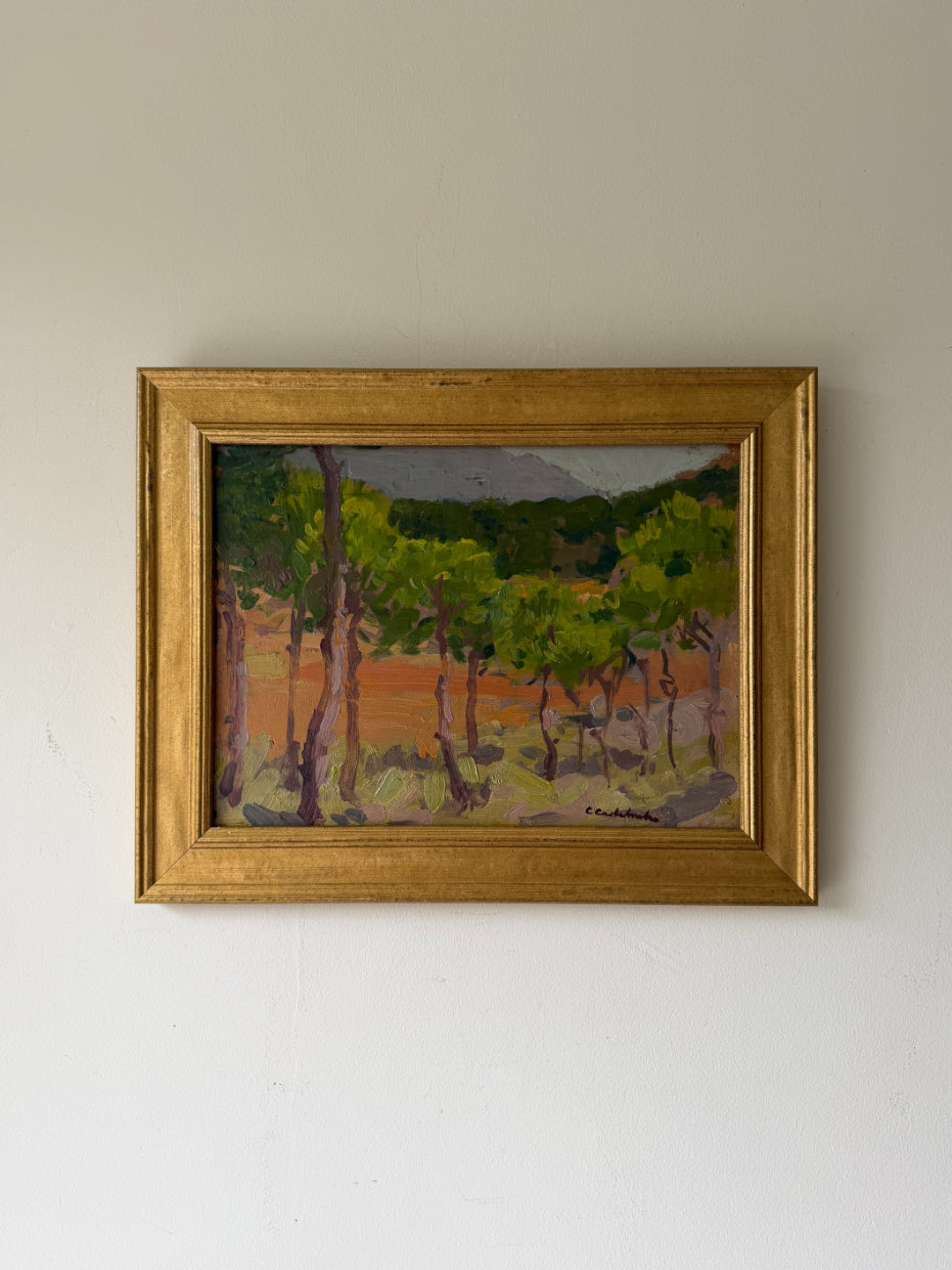 A framed vintage oil landscape painting featuring green trees and a warm orange-terracotta field. Available at Hue.