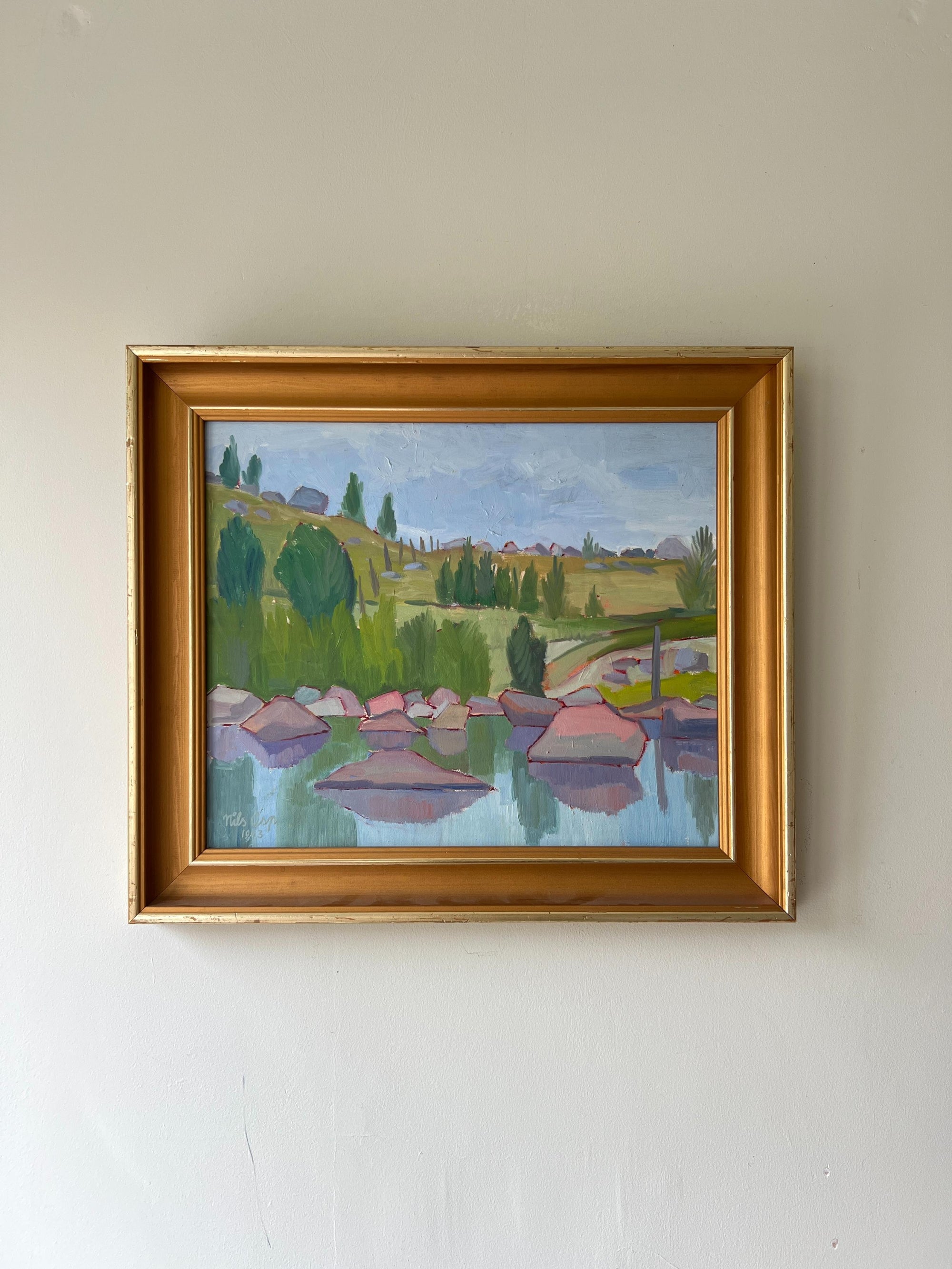 A framed vintage oil painting of a lake shore with pink rocks and landscape beyond. Available at Hue. 