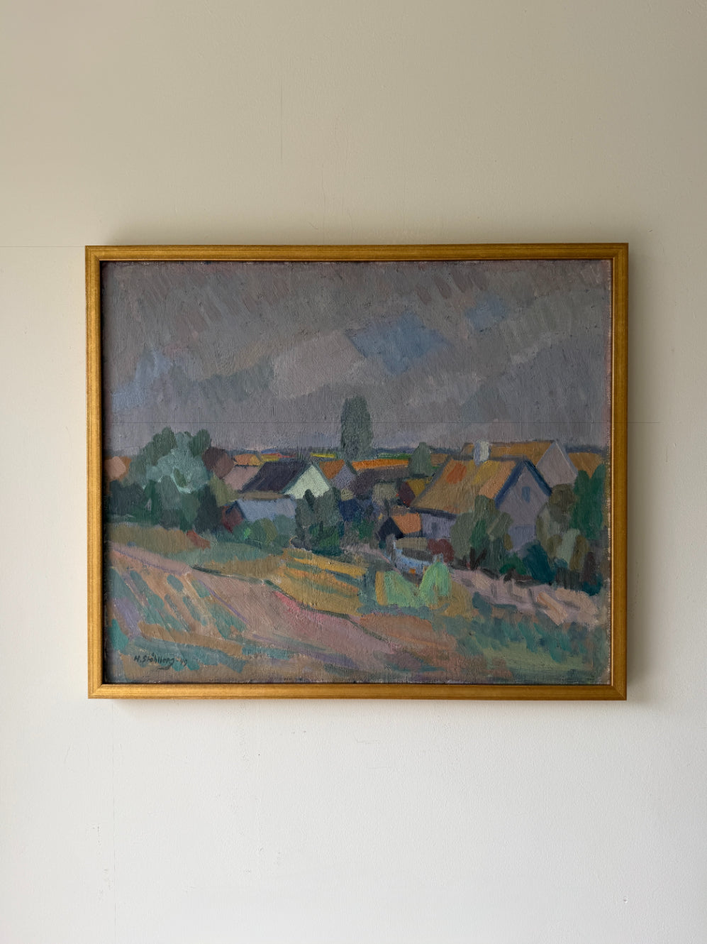 A framed vintage landscape oil painting of a small village with houses, trees, and a cloudy sky. Available at Hue. 