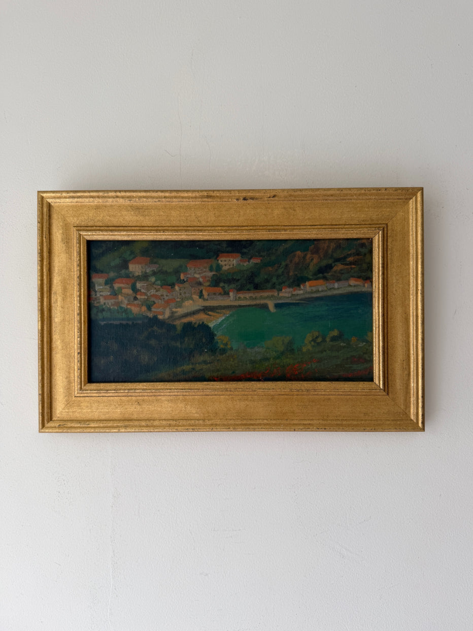 A framed vintage landscape oil painting depicting a coastal town with turquoise water. Available at Hue. 