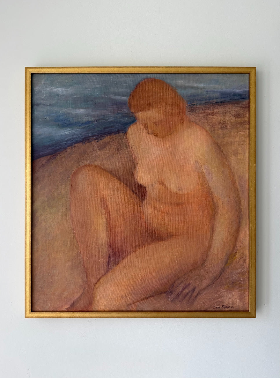 A framed vintage painting of a seated nude woman on a beach, with blue water in the background and a gold frame. Available at Hue.