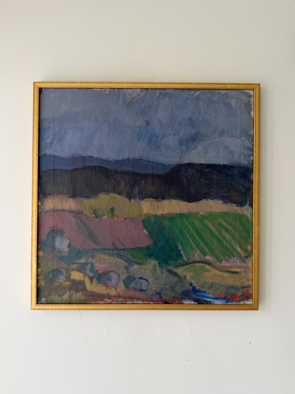 A framed vintage landscape oil painting featuring green fields, dark hills, and a stormy blue-gray sky, enclosed in a thin gold frame. Available at Hue.