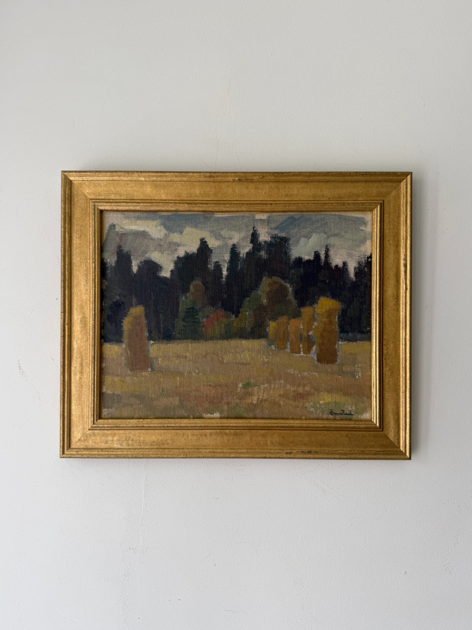 A framed vintage landscape painting. The painting depicts a field with hayricks under a dark forest and cloudy sky, enclosed in a wide gold frame. Available at Hue.
