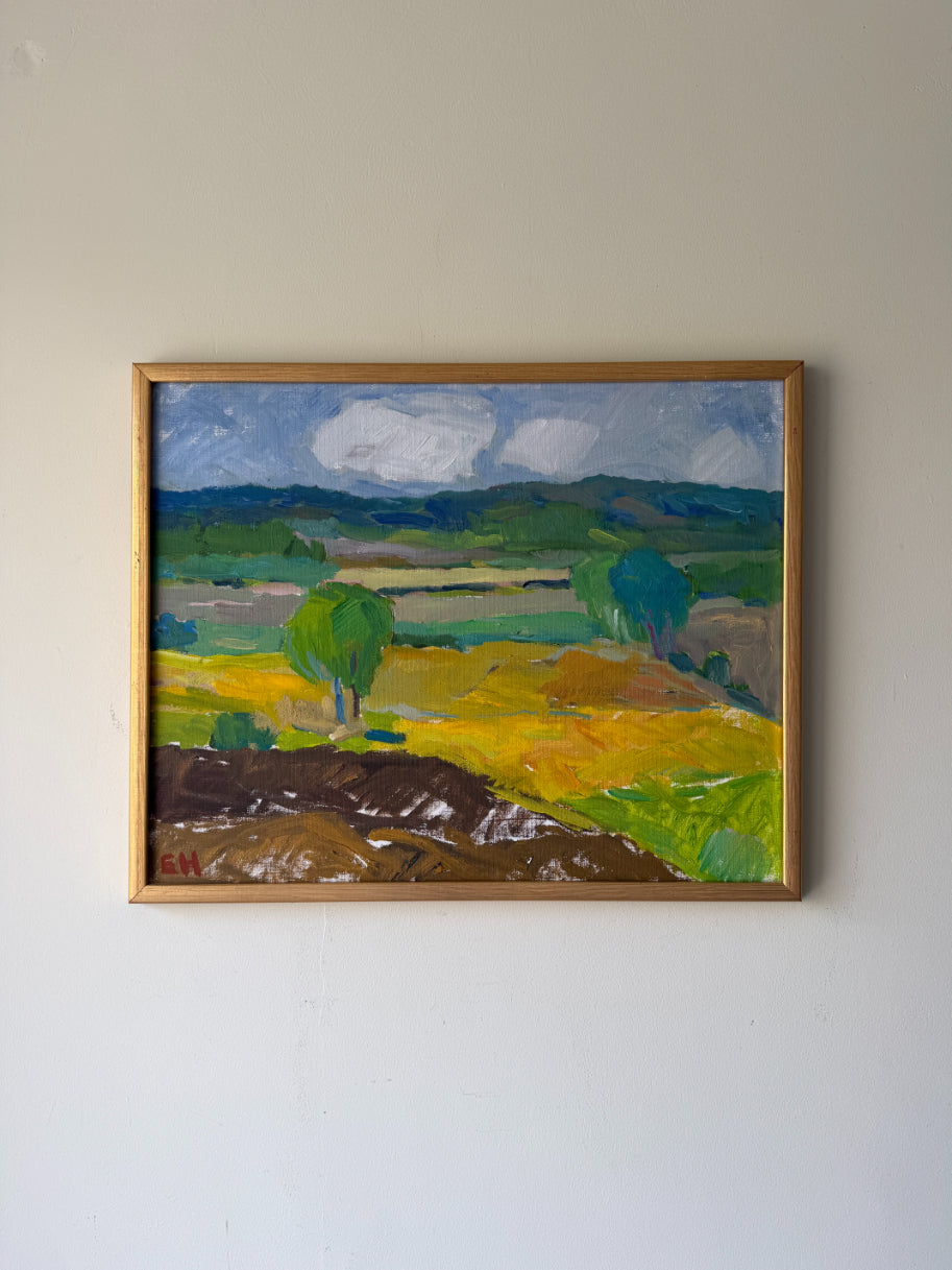 A framed vintage landscape oil painting. The painting depicts rolling green hills, yellow fields, a brown foreground, and a blue-gray cloudy sky. Available at Hue. 